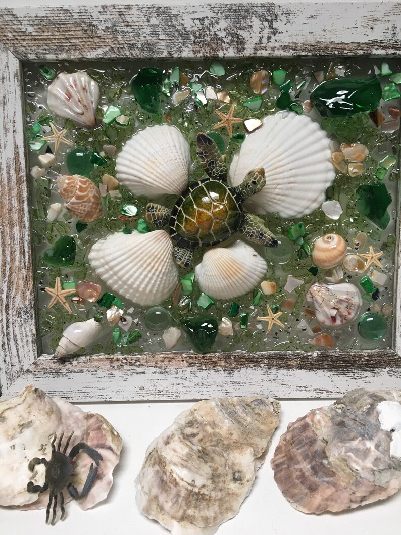Puede incluir: Una pieza de arte de resina enmarcada que presenta una tortuga marina verde rodeada de conchas marinas y piezas de vidrio verde. El marco es blanco con un acabado desgastado.