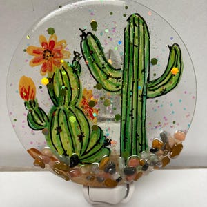 May include: A nightlight with a clear glass dome featuring a hand-painted design of two cacti with green, yellow, and orange accents. The cacti are surrounded by small brown and white stones. The nightlight is plugged into an electrical outlet.