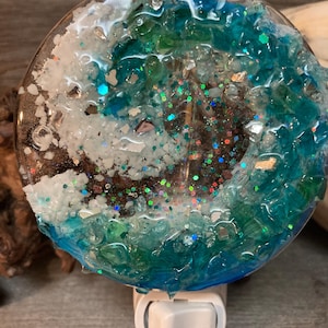May include: A blue and green resin nightlight with glitter and white accents. The nightlight is shaped like a wave and has a white electrical plug.