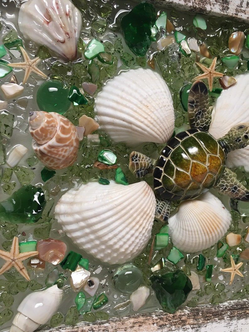 Puede incluir: Una pieza de arte de resina con una tortuga marina verde, conchas marinas y piezas de vidrio verde. La tortuga est&aacute; en el centro de la pieza y est&aacute; rodeada de conchas marinas y piezas de vidrio.