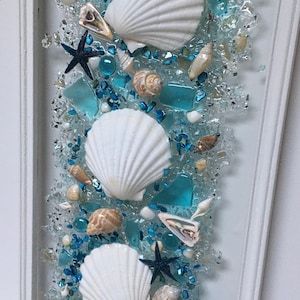 May include: A framed wall art piece featuring a white frame and a blue and white seashell design. The design includes three white seashells, blue glass pieces, and other seashells.