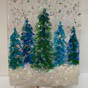 May include: A white nightlight with a clear glass front featuring a winter scene with five blue and green trees painted on a white background with white glitter.