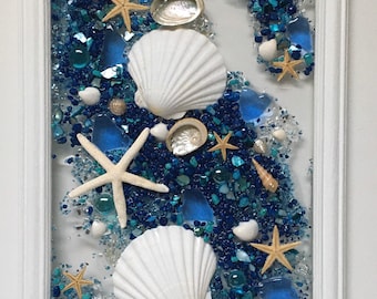 May include: A white framed artwork featuring a seashell and starfish design. The artwork is filled with blue and turquoise glass pieces and white seashells.