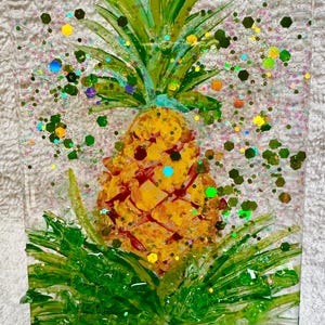 May include: A decorative night light featuring a yellow pineapple design with green leaves and glitter accents. The pineapple is set against a clear background with a white plug at the bottom. The artwork is encased in a rectangular shape.
