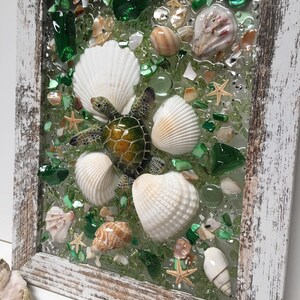 Puede incluir: Una pieza de arte de mosaico enmarcada que presenta una tortuga marina verde rodeada de conchas blancas y piezas de vidrio verde. El fondo es de color verde claro.