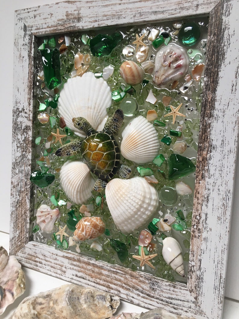 Puede incluir: Una pieza de arte de mosaico enmarcada que presenta una tortuga marina verde rodeada de conchas blancas y piezas de vidrio verde. El fondo es de color verde claro.