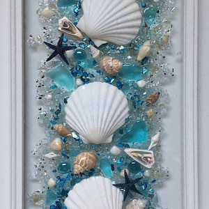 May include: A white framed wall art piece with a vertical arrangement of three white seashells, blue and white glass beads, and small seashells. The background is a light blue colour.