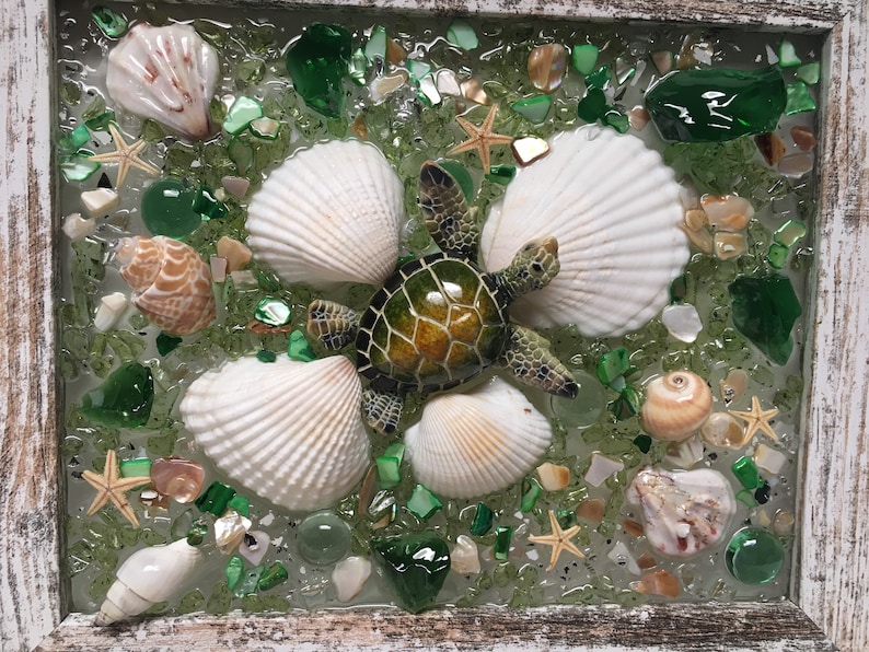 Puede incluir: Una pieza de arte de resina enmarcada que presenta una tortuga marina verde rodeada de conchas blancas y piezas de vidrio verde e iridiscente.