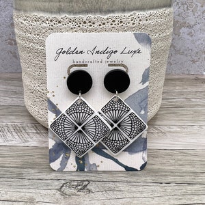 Puede incluir: Un par de pendientes geométricos con estampado en blanco y negro con postes negros. Los pendientes se muestran en una tarjeta con el texto "Golden Indigo Luxe handcrafted jewelry".