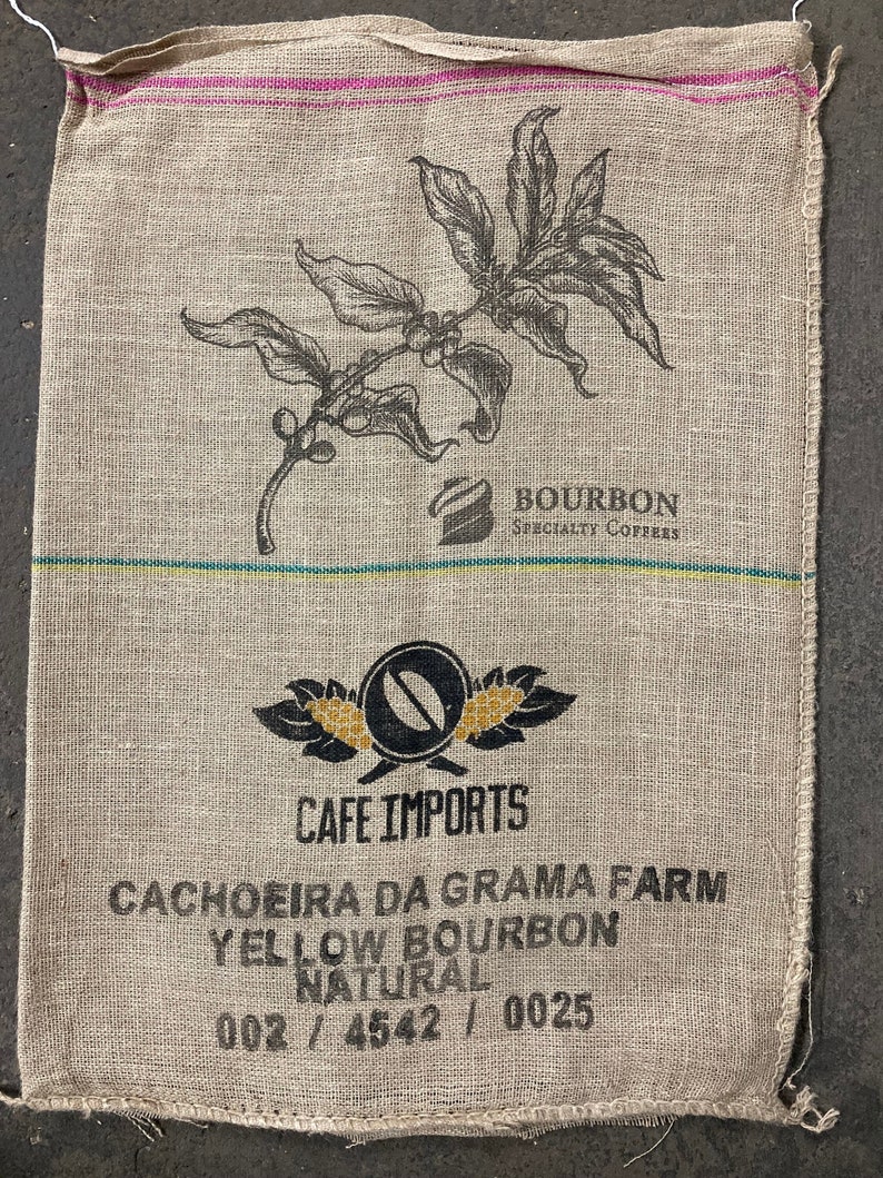 May include: A burlap coffee bag with a black and yellow logo that says "Cafe Imports". The bag also has a black and white illustration of a coffee plant and the text "Bourbon Specialty Coffees", "Cachoeira da Grama Farm", "Yellow Bourbon Natural", "002 / 4542 / 0025".