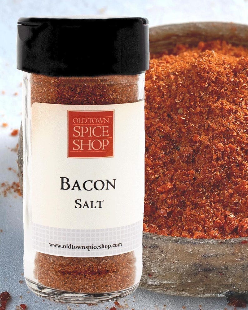 May include: A clear glass spice jar with a black lid containing reddish-brown bacon salt. The white label features the red Old Town Spice Shop logo, the text Bacon Salt, and the website address www.oldtownspiceshop.com in black lettering.