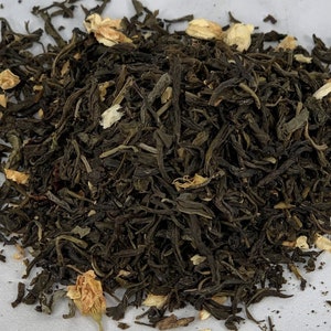 May include: A close-up of a pile of loose leaf green tea with white jasmine flower petals. The tea is a dark green color and the petals are a light white.
