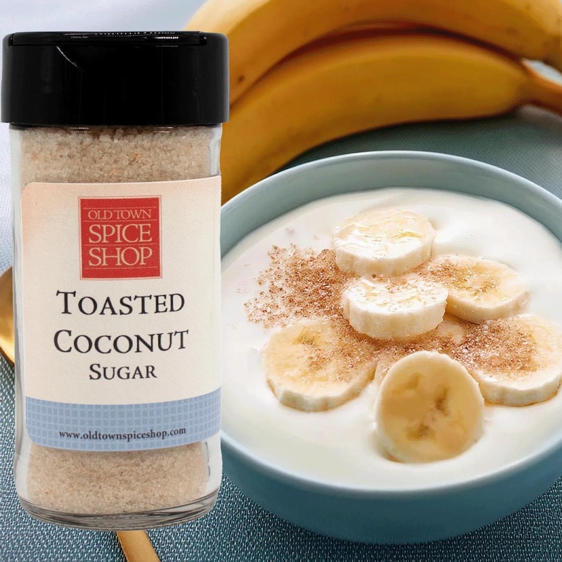 May include: A glass jar of Old Town Spice Shop Toasted Coconut Sugar sits beside a bowl of yogurt topped with banana slices and sugar. Two bananas are in the background. The jar has a black lid and a label with product details.