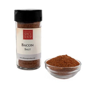 Puede incluir: Un tarro de vidrio de sal de tocino Old Town Spice Shop. El tarro tiene una tapa negra y una etiqueta blanca con texto rojo que dice "Old Town Spice Shop" y "Bacon Salt". El tarro est&aacute; lleno de un condimento marr&oacute;n granulado.
