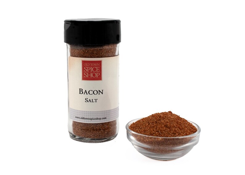 Puede incluir: Un tarro de vidrio de sal de tocino Old Town Spice Shop. El tarro tiene una tapa negra y una etiqueta blanca con texto rojo que dice "Old Town Spice Shop" y "Bacon Salt". El tarro est&aacute; lleno de un condimento marr&oacute;n granulado.