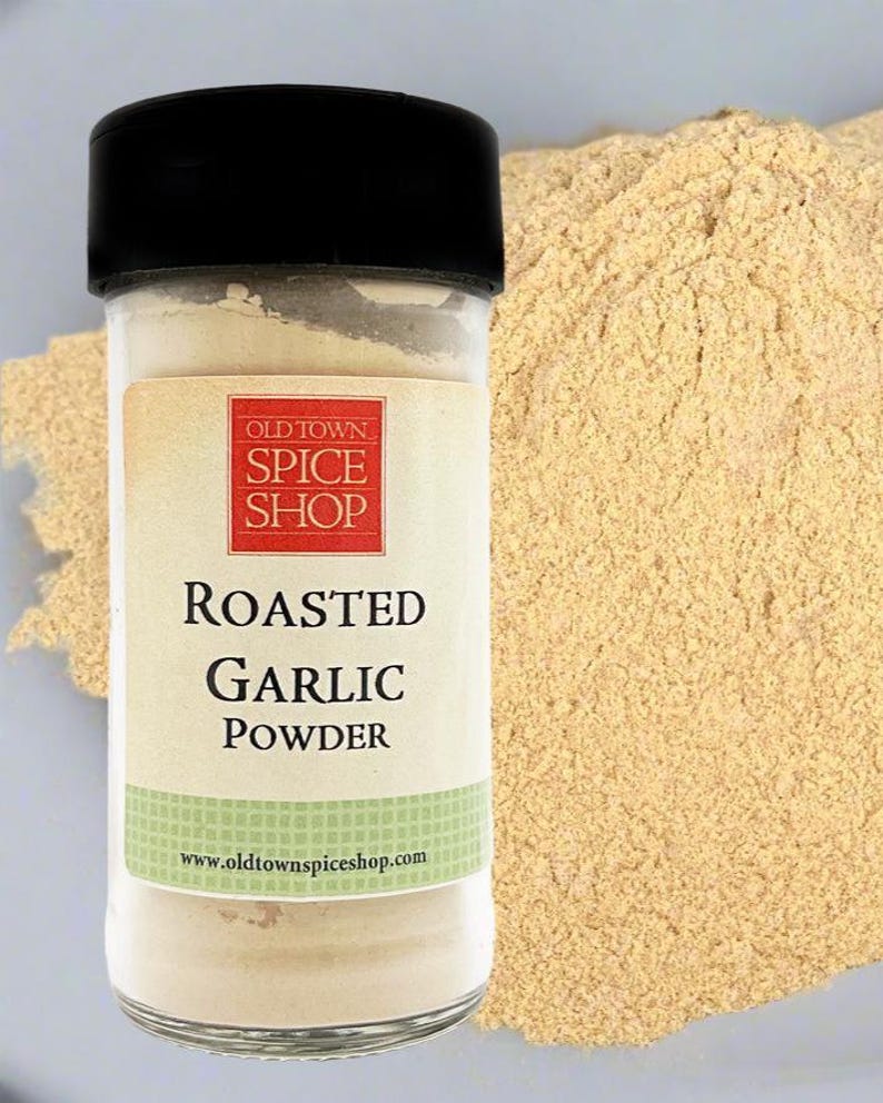 Puede incluir: Un frasco de especias de vidrio transparente con tapa negra que contiene ajo en polvo tostado. La etiqueta presenta un cuadrado rojo con el texto OLD TOWN SPICE SHOP, seguido de las palabras ROASTED GARLIC POWDER y el sitio www.oldtownspiceshop.com.