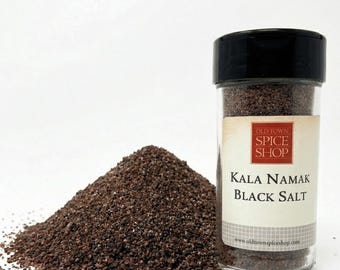 May include: A clear plastic spice jar with a black lid containing dark brown Kala Namak black salt. The label reads Old Town Spice Shop, Kala Namak Black Salt, and includes the website address. A pile of the same dark salt sits beside the jar.