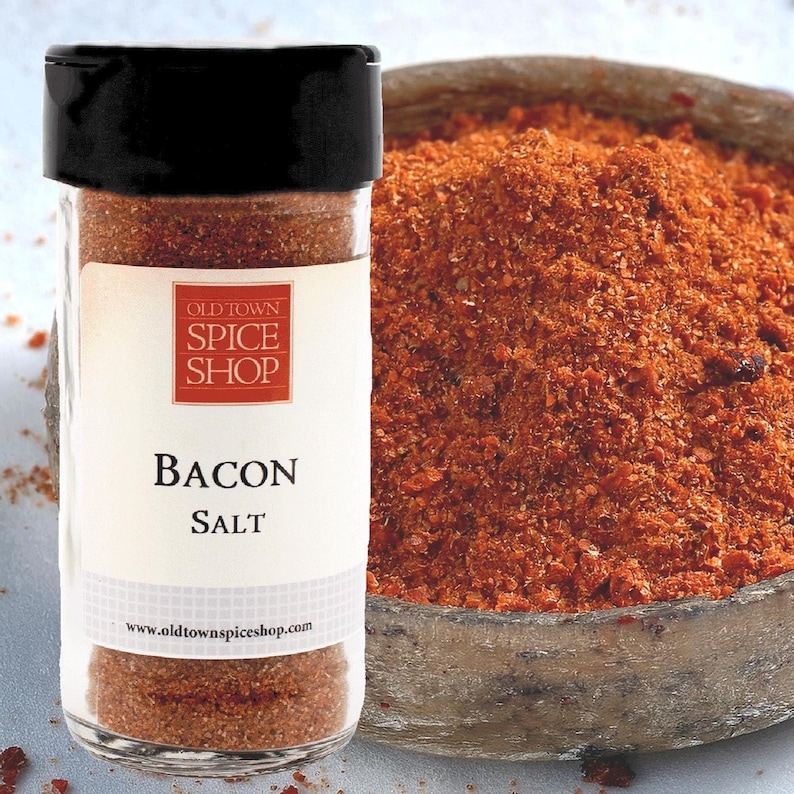 Puede incluir: Un tarro de vidrio lleno de sal de tocino marr&oacute;n y roja. El tarro tiene una tapa negra y una etiqueta blanca que dice "Old Town Spice Shop Bacon Salt" y "www.oldtownspiceshop.com".