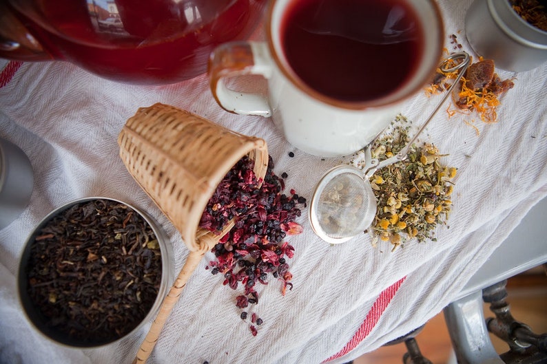May include: A close-up of a white tea strainer filled with dried berries, a white teacup with red tea, a small tin of loose leaf tea, and a silver tea strainer filled with dried herbs. The items are arranged on a white linen tablecloth with red stripes.
