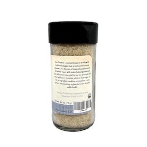 May include: A clear glass jar with a black lid containing light brown sugar. The label states "Our Toasted Coconut Sugar" and details ingredients and uses. Net weight is 73 grams.