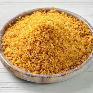 May include: A round, shallow bowl with a mottled grey and brown finish is filled to the brim with a mound of coarse, golden-yellow flakes. The texture of the contents appears dry and granular, with varying sizes of small, irregular pieces.