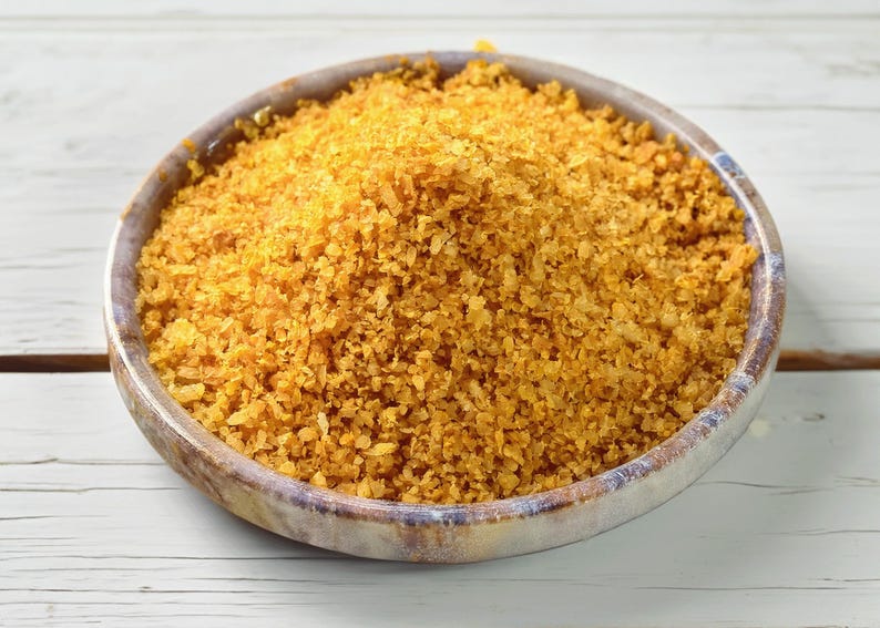 May include: A round, shallow bowl with a mottled grey and brown finish is filled to the brim with a mound of coarse, golden-yellow flakes. The texture of the contents appears dry and granular, with varying sizes of small, irregular pieces.