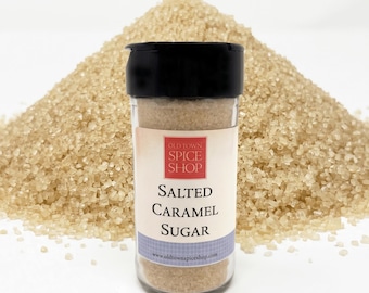 Könnte beinhalten: Ein klares Glas mit schwarzem Deckel, beschriftet mit "Salted Caramel Sugar" von Old Town Spice Shop, steht vor einem Haufen brauner Zuckerkristalle. Das Etikett hat ein rot-weißes Logo und einen blau-weiß gemusterten Streifen.