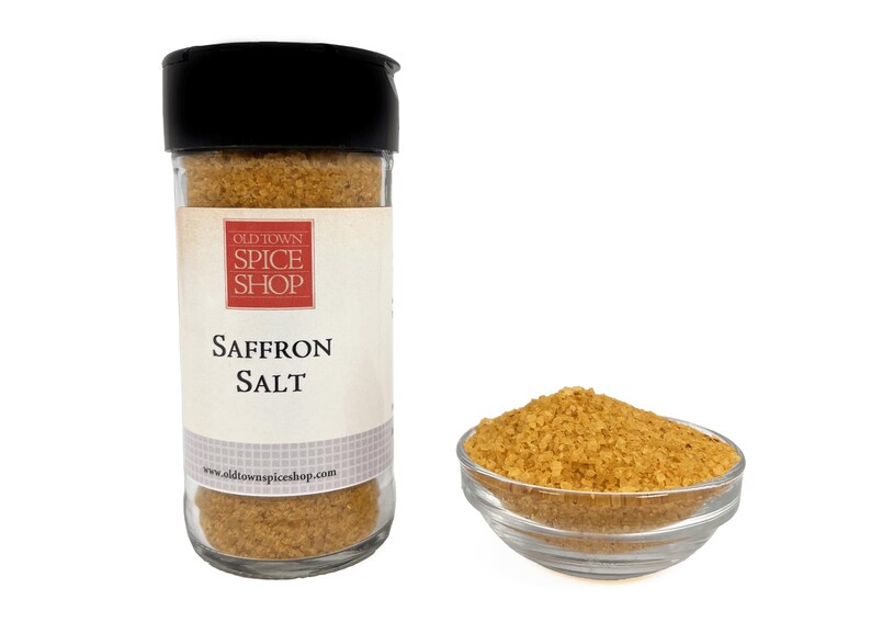 May include: A clear glass spice jar with a black lid containing golden-yellow saffron salt. The label features the text Old Town Spice Shop and Saffron Salt. Next to the jar is a small clear glass bowl filled with the same golden-yellow saffron salt.
