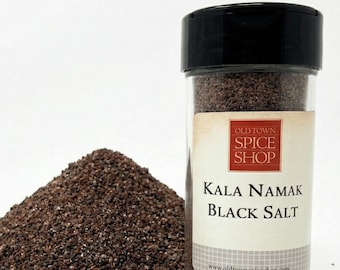 May include: A clear glass spice jar with a black lid containing dark brown, coarse-grained Kala Namak black salt. The label features the text Old Town Spice Shop, Kala Namak Black Salt, and www.oldtownspiceshop.com. A pile of the salt sits beside it.