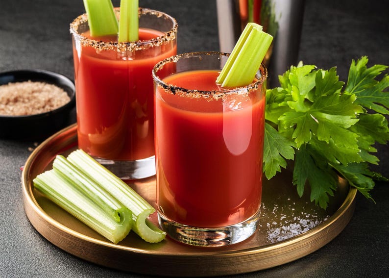 Puede incluir: Dos vasos de zumo de tomate rojo con tallos de apio y un borde salado. Los vasos est&aacute;n en una bandeja dorada con una guarnici&oacute;n verde de hojas.