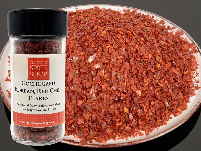 May include: A glass jar filled with red chili flakes labeled "Gochugaru Korean, Red Chili Flakes". The label also states "Warm and fruity in flavor with a heat that ranges from mild to hot" and includes the website "www.oldtownspiceshop.com".