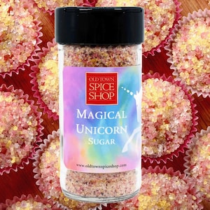 May include: A glass jar of pink and yellow sugar with the label "Old Town Spice Shop Magical Unicorn Sugar" and the website www.oldtownspiceshop.com. The jar is surrounded by pink and yellow cupcake liners filled with pink and yellow sugar.