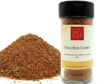 May include: A clear plastic spice jar with a black lid containing Thai Red Curry seasoning, featuring a white label with red text reading Old Town Spice Shop and Thai Red Curry. A pile of the reddish-brown spice blend sits next to the jar.