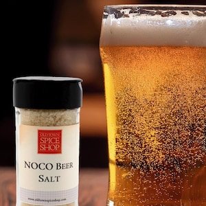 May include: A clear glass spice shaker with a black lid containing coarse, light-colored grains. The label reads Old Town Spice Shop, Noco Beer Salt, and www.oldtownspiceshop.com. Beside the shaker is a tall glass filled with golden beer.