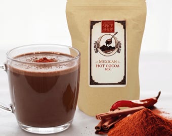 Może przedstawiać: Przezroczysty szklany kubek wypełniony ciemnobrązowym gorącym kakao z posypką, obok beżowej torebki z etykietą Old Town Spice Shop Mexican Hot Cocoa Mix, czerwonej papryczki chili, lasek cynamonu i kopczyka czerwonobrązowego proszku z przyprawami.