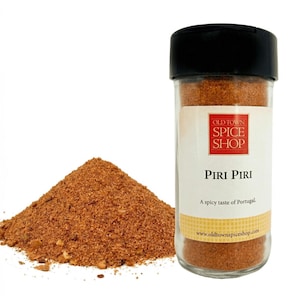 May include: A clear glass jar of Piri Piri spice from Old Town Spice Shop with a black lid. A pile of the reddish-brown spice is next to the jar. The label says "A spicy taste of Portugal."