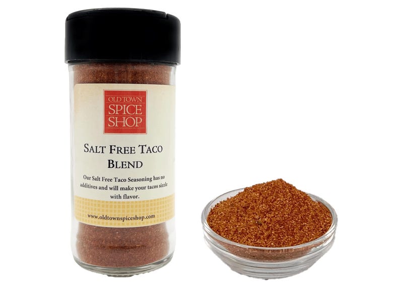 Pu&ograve; includere: Un barattolo di vetro di miscela di condimento per tacos senza sale con un'etichetta marrone che recita "Old Town Spice Shop Salt Free Taco Blend". L'etichetta include anche l'indirizzo del sito web "www.oldtownspiceshop.com". Una piccola ciotola di condimento &egrave; mostrata accanto al barattolo.