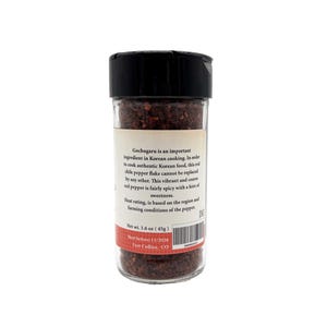 May include: A glass jar filled with red gochugaru Korean chili pepper flakes. The jar has a black lid and a white label with the text "Gochugaru is an important ingredient in Korean cooking. In order to cook authentic Korean food, this red chili pepper flake cannot be replaced by any other. This vibrant and coarse red pepper is fairly spicy with a hint of sweetness. Heat rating is based on the region and farming conditions of the pepper. Net wt. 1.4 oz (45g). Best before 11/1/2024. Fort Collins, CO."