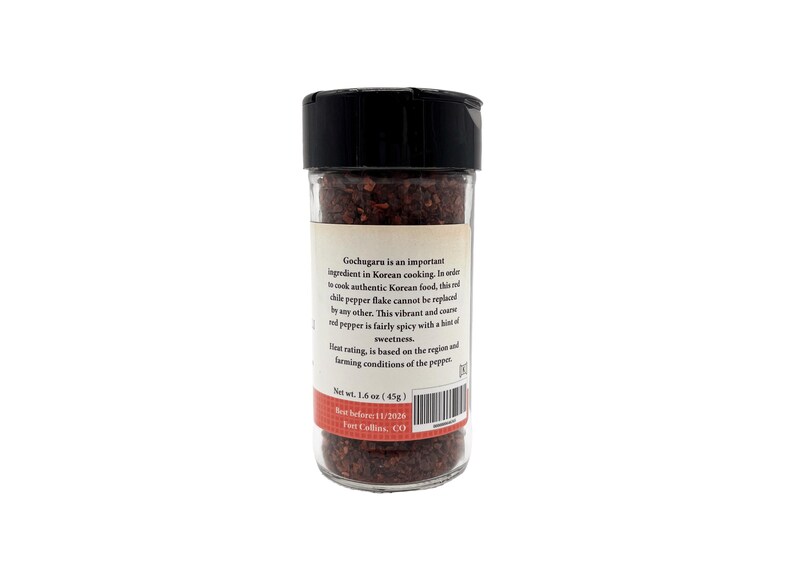 May include: A glass jar filled with red gochugaru Korean chili pepper flakes. The jar has a black lid and a white label with the text "Gochugaru is an important ingredient in Korean cooking. In order to cook authentic Korean food, this red chili pepper flake cannot be replaced by any other. This vibrant and coarse red pepper is fairly spicy with a hint of sweetness. Heat rating is based on the region and farming conditions of the pepper. Net wt. 1.4 oz (45g). Best before 11/1/2024. Fort Collins, CO."