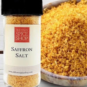 May include: A clear glass jar with a black lid containing golden-yellow saffron salt. The label features a red square with the text OLD TOWN SPICE SHOP and the words SAFFRON SALT below it. A bowl filled with the same yellow salt sits beside the jar.