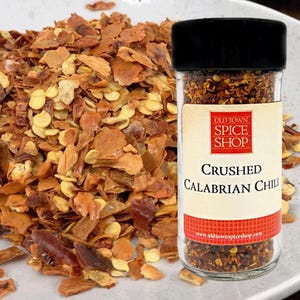 May include: A clear glass spice jar with a black lid containing crushed Calabrian chili flakes. The label features the text Old Town Spice Shop, Crushed Calabrian Chili, and the website address. A pile of the same chili flakes sits beside it.