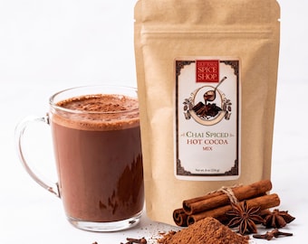 Peut inclure: Un mug en verre transparent rempli de chocolat chaud brun, à côté d'un sachet en papier beige étiqueté Old Town Spice Shop Chai Spiced Hot Cocoa Mix, 226 g, avec des bâtons de cannelle, de l'anis étoilé et de la poudre de cacao.