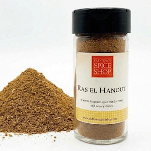 May include: A clear glass spice jar with a black lid containing brown Ras El Hanout spice mix. The label reads Old Town Spice Shop, Ras El Hanout, A warm, fragrant spice mix for sweet and savory dishes, and www.oldtownspiceshop.com. A pile of the spice sits beside the jar.