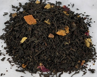 May include: Close-up of loose leaf tea with dark brown leaves and pieces of dried fruit and rose petals. The tea is scattered on a white surface, showcasing the texture and variety of ingredients.