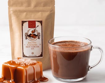 Może przedstawiać: Brązowa papierowa torebka z etykietą Old Town Spice Shop Salted Caramel Hot Cocoa Mix, waga netto 227g, obok przezroczystego szklanego kubka wypełnionego ciemnobrązowym gorącym kakao oraz kostką karmelu z różową solą morską.