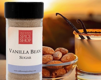 Könnte beinhalten: Ein Glasgefäß mit Vanille-Zucker und schwarzem Deckel. Das Etikett lautet "Old Town Spice Shop" und "Vanilla Bean Sugar". Das Gefäß steht auf einem Holztisch mit einem Glas Vanille-Getränk und einer Schale mit Vanille-Mandeln.
