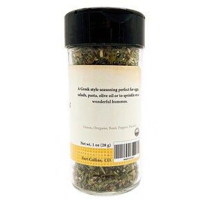 May include: A clear plastic spice jar with a black lid containing dried green herbs. The label reads: A Greek style seasoning perfect for eggs, salads, pasta, olive oil or to sprinkle on a wonderful hummus. Onion, Oregano, Basil, Pepper, Parsley. Net wt. 1 oz (28 g). Fort Collins, CO.
