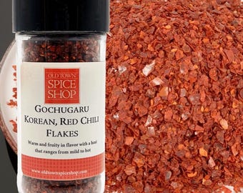 May include: A clear plastic spice jar with a black lid containing Gochugaru Korean red chili flakes. The label reads Old Town Spice Shop, Gochugaru Korean, Red Chili Flakes, and includes text about flavor. A pile of red chili flakes sits behind the jar.