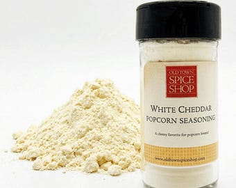 Puede incluir: Un frasco de especias de plástico transparente con tapa negra que contiene condimento de cheddar blanco para palomitas. La etiqueta muestra el texto Old Town Spice Shop, White Cheddar Popcorn Seasoning y una URL. Un montón de condimento está al lado.