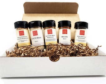 May include: A white cardboard box with brown paper packing peanuts inside. Five glass jars with black lids are sitting on top of the packing peanuts. The jars contain different spice blends, including "Bacon Cheddar Popcorn Seasoning", "Noco Beer Salt", "Backyard Brew Marinade", "Bacon Salt", and "Beer Popcorn Seasoning".